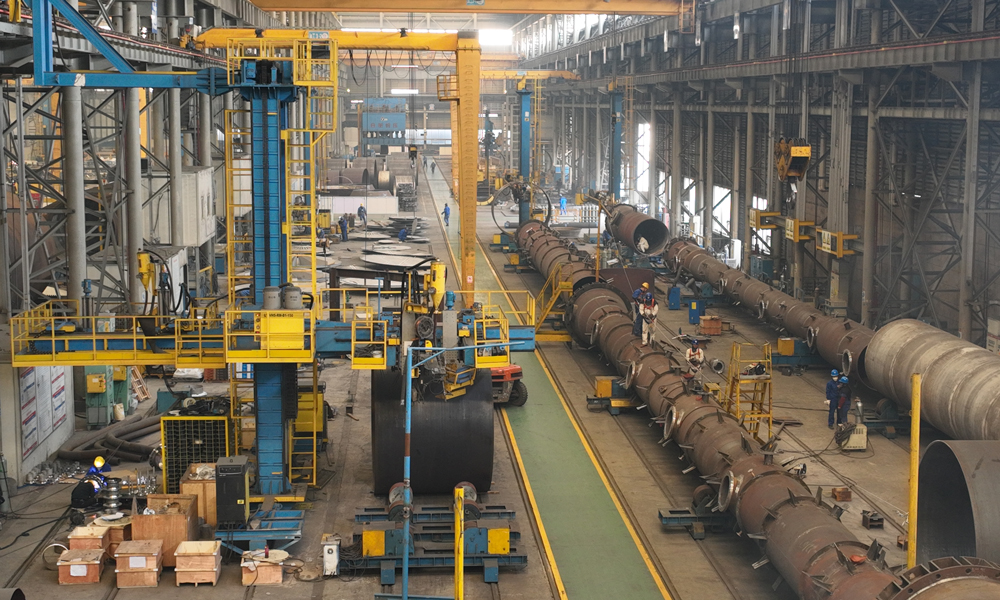 在內蒙古圖克工業區的制造現場，上百噸的“龐然大物”被拆解成一塊塊“積木”：異型錐體被分片壓制、組裝；數十噸鋼板一遍遍“翻身”，10米立車在鋼花中精雕細琢……與此同時，1500多公里外的南京車間內，工人們為巨大的“滾筒”噴上白漆，設備內焊花飛濺。這是中建安裝旗下中建五洲“現場+工廠”兩線制造模式在寶豐項目的再度應用。憑借著全國領先的大型塔器、反應器現場制造水平，項目團隊完成了內蒙古寶豐項目反應、換熱、分離及儲存容器等91臺重要設備制造與安裝。